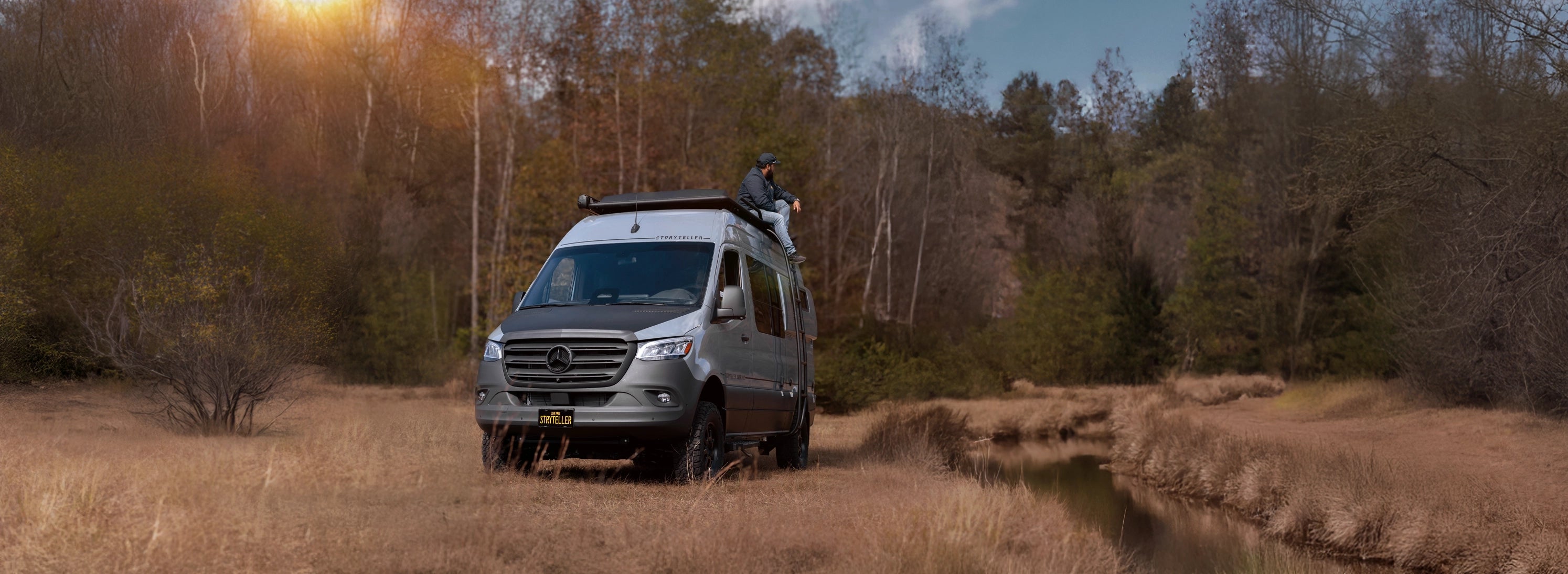Van parked near a forest with a person on top, surrounded by trees and nature.