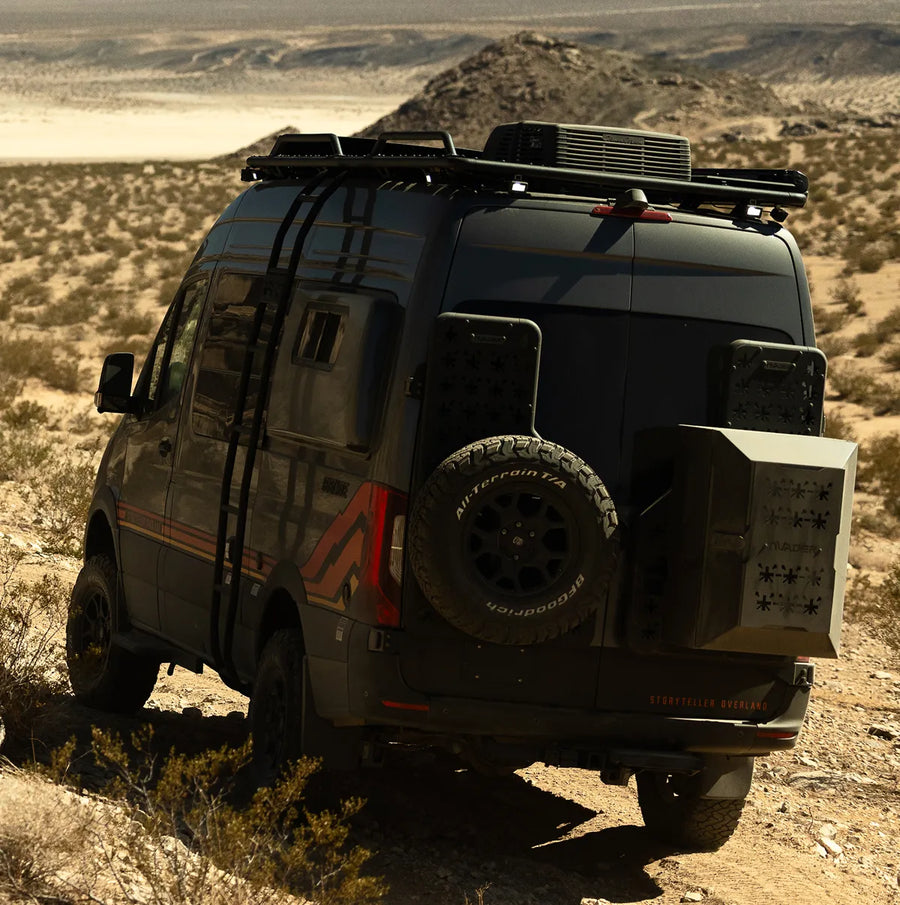 Black Ford Transit 4x4 camper van offroading in desert, equipped for overlanding adventure with roof rack and spare tire