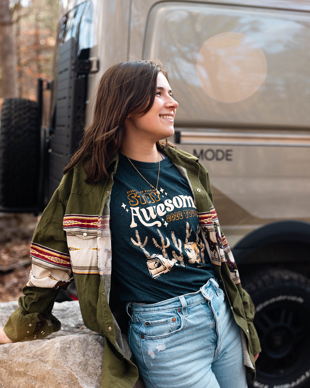 Smiling woman in front of a MODE4x4 adventure van, wearing Stay Awesome tour shirt, outdoors